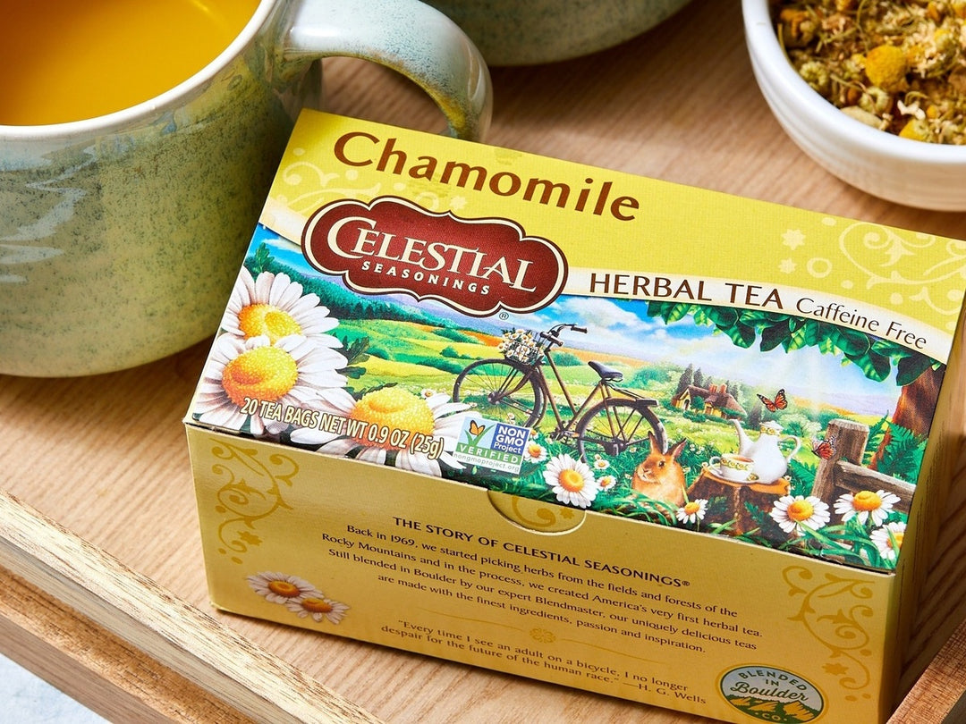 Celestial Seasonings Chamomile Herbal Tea box shown on a wooden surface with a mug of tea and a bowl of dried chamomile flowers nearby.