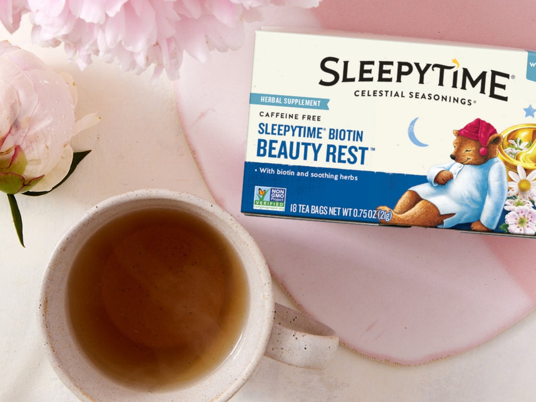 A box of Sleepytime Caffeine Free Herbal Supplement tea by Celestial Seasonings is displayed next to a cup of brewed tea on a pink tray with pink flowers.