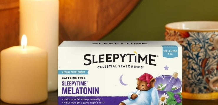 A box of Sleepytime Melatonin herbal supplement tea by Celestial Seasonings, styled on a wooden surface with a lit candle on the left and a patterned mug on the right.