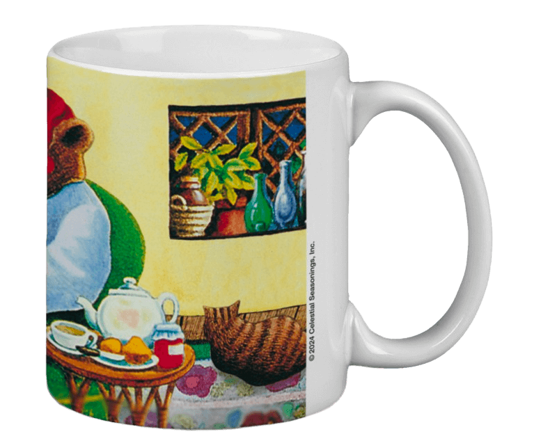 White mug featuring a cozy illustration of a bear in a red hat sitting indoors. A table holds a teapot, cup, and jam. A window shows plants and bottles.