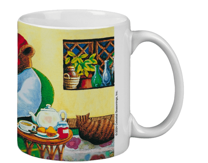 White mug featuring a cozy illustration of a bear in a red hat sitting indoors. A table holds a teapot, cup, and jam. A window shows plants and bottles.