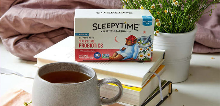 A box of Sleepytide Celestial Seasonings Sleepytime Probiotics tea is displayed on a stack of books, with a gray ceramic mug of tea in the foreground and a potted plant in the background.